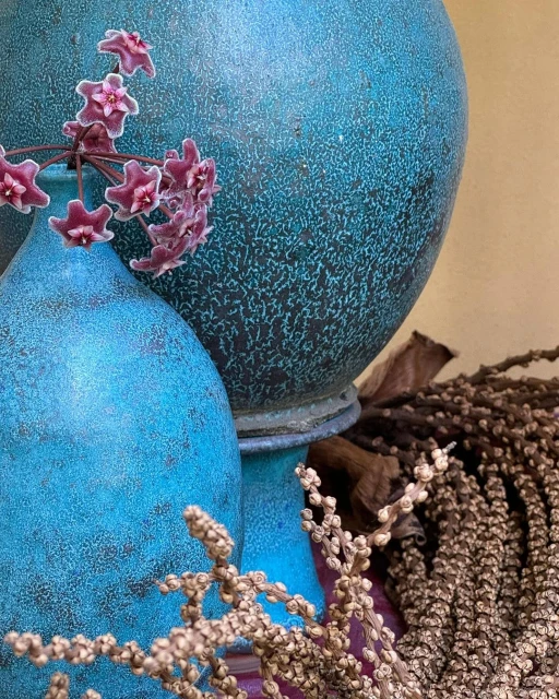 Close-up of teal blue textured pottery vases with a small arrangement of burgundy star-shaped flowers and dried botanical elements.