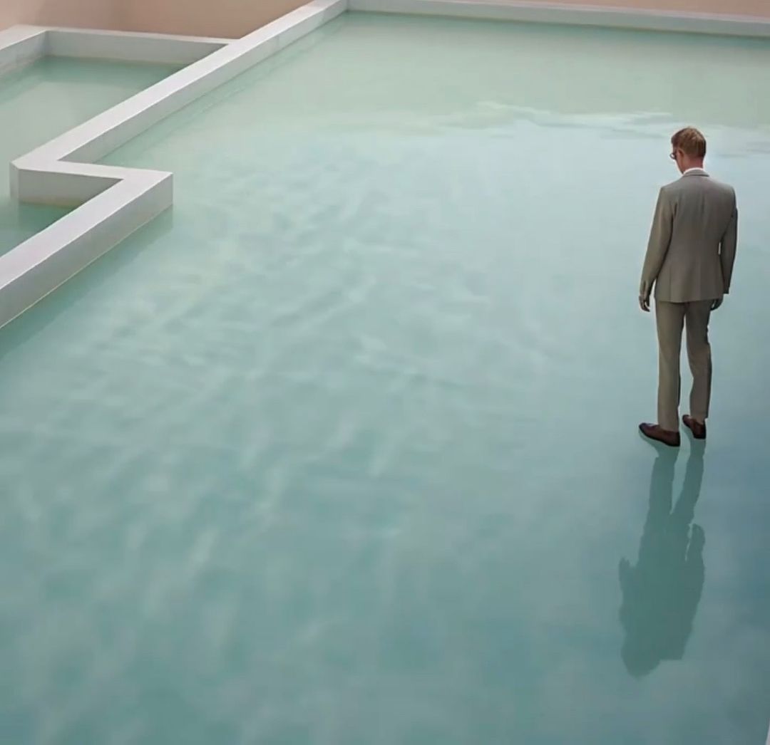 Man in a suit standing on a flooded floor with a zig-zag barrier, facing away from the camera, creating a surreal, abstract scene.