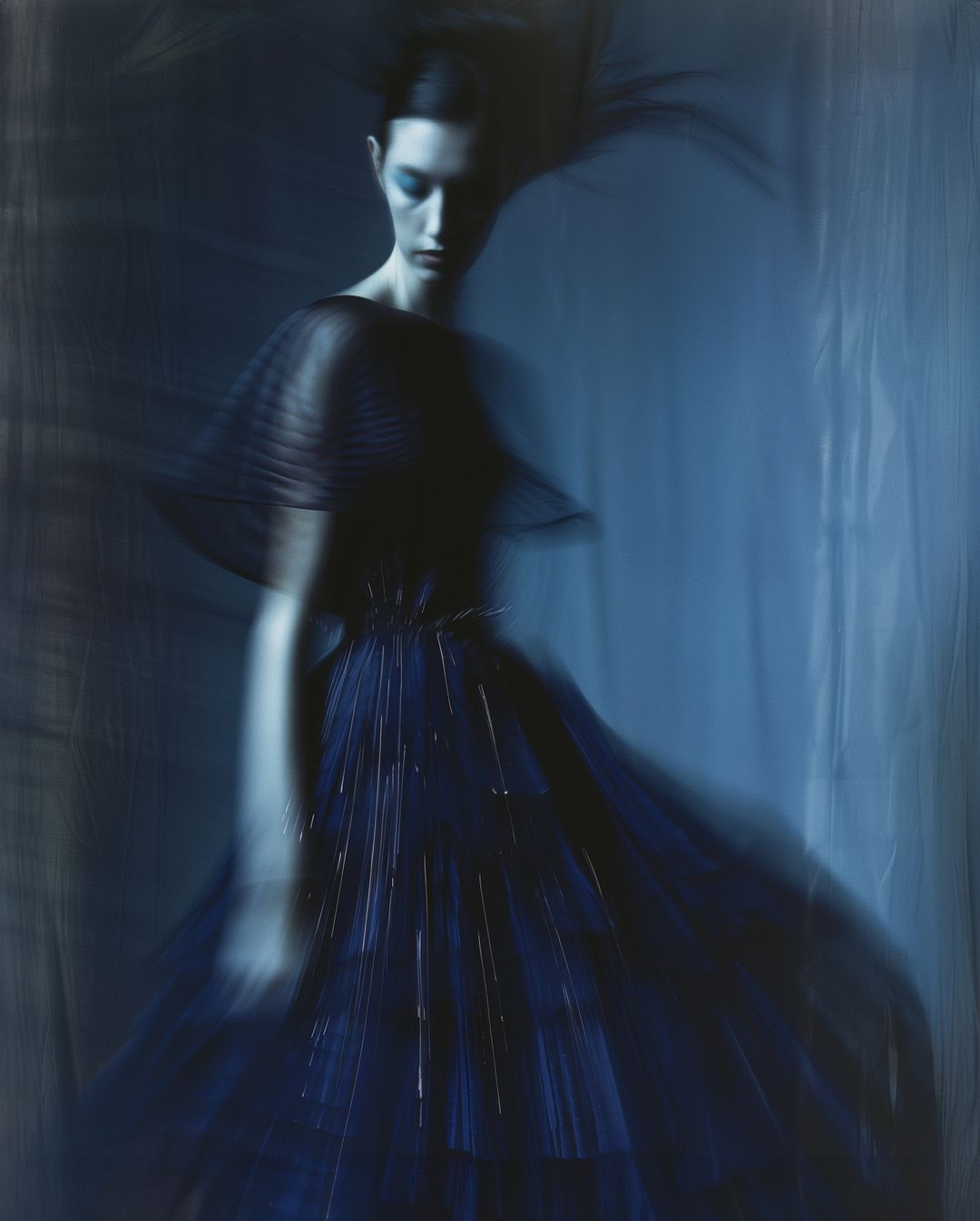 An adult female fashion model poses in a dark blue dress with flowing layers in a photo studio with artistic blur and low-key lighting.