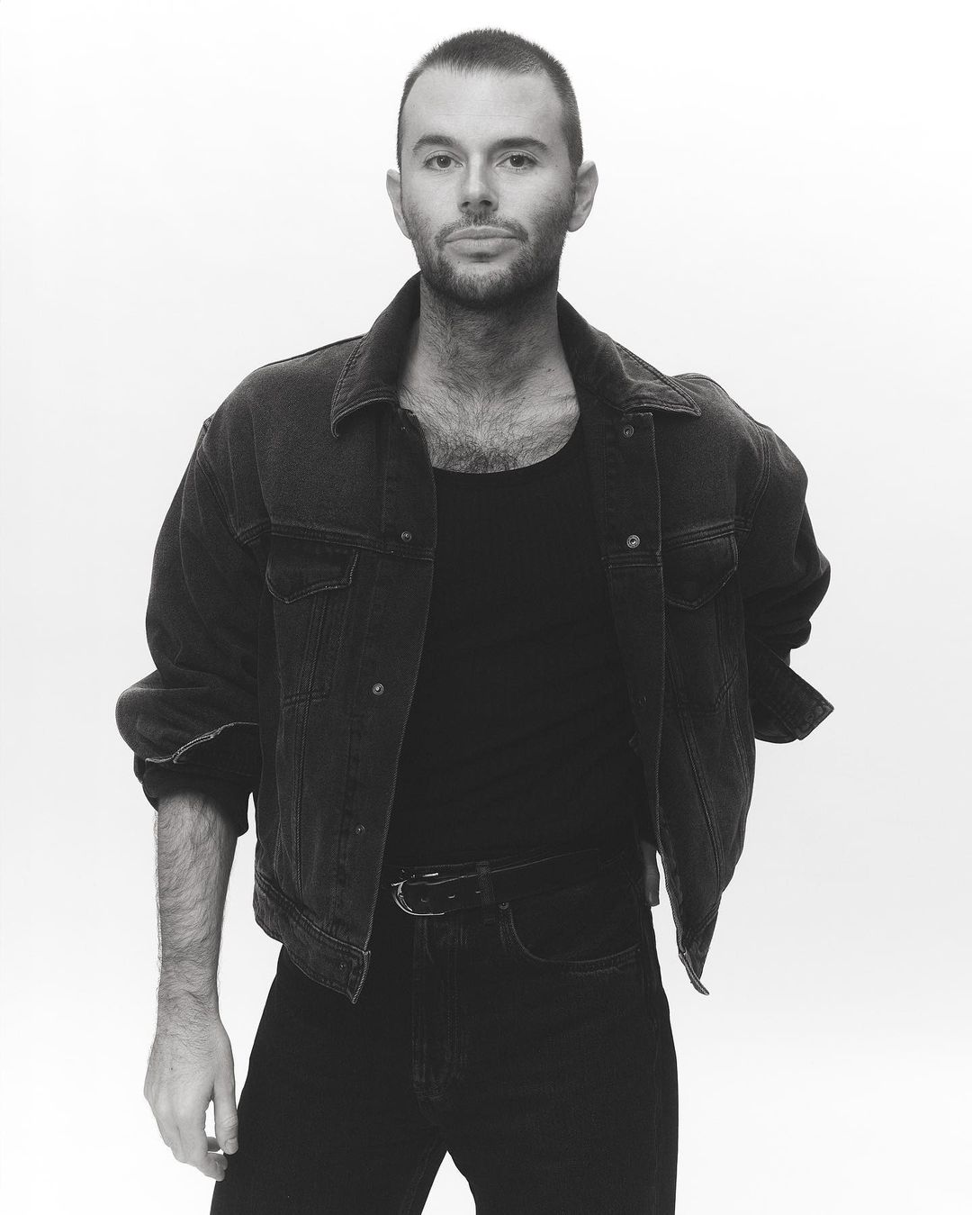 Black and white portrait of a man in a denim jacket, black shirt, and black jeans against a white backdrop in a photo studio.