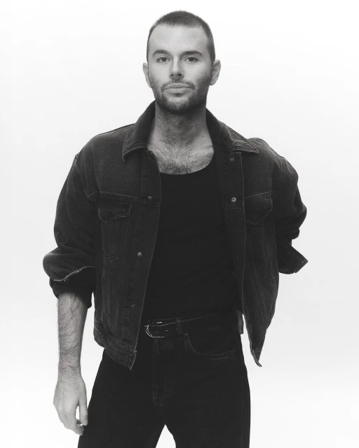 Black and white portrait of a man in a denim jacket, black shirt, and black jeans against a white backdrop in a photo studio.