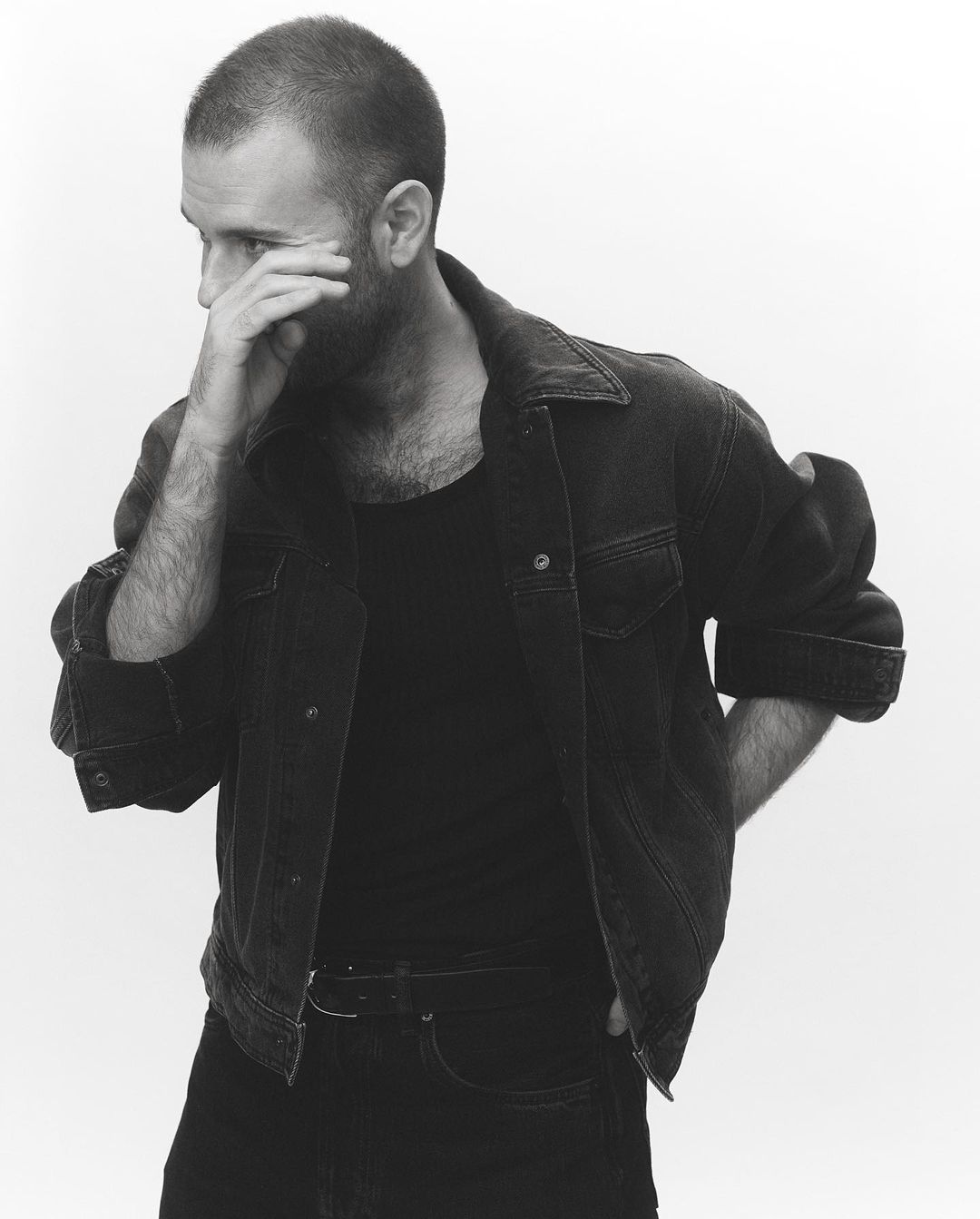 Monochrome portrait of a man wearing a dark denim jacket and t-shirt with his hand near his face against a white background.