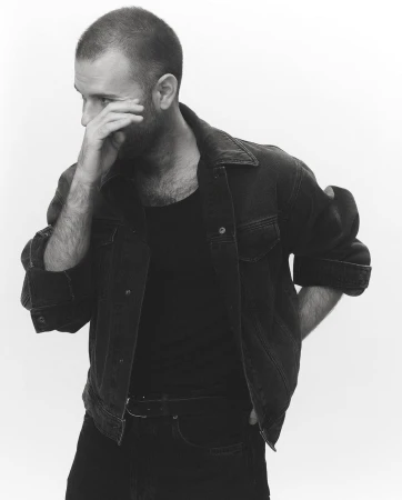 Monochrome portrait of a man wearing a dark denim jacket and t-shirt with his hand near his face against a white background.