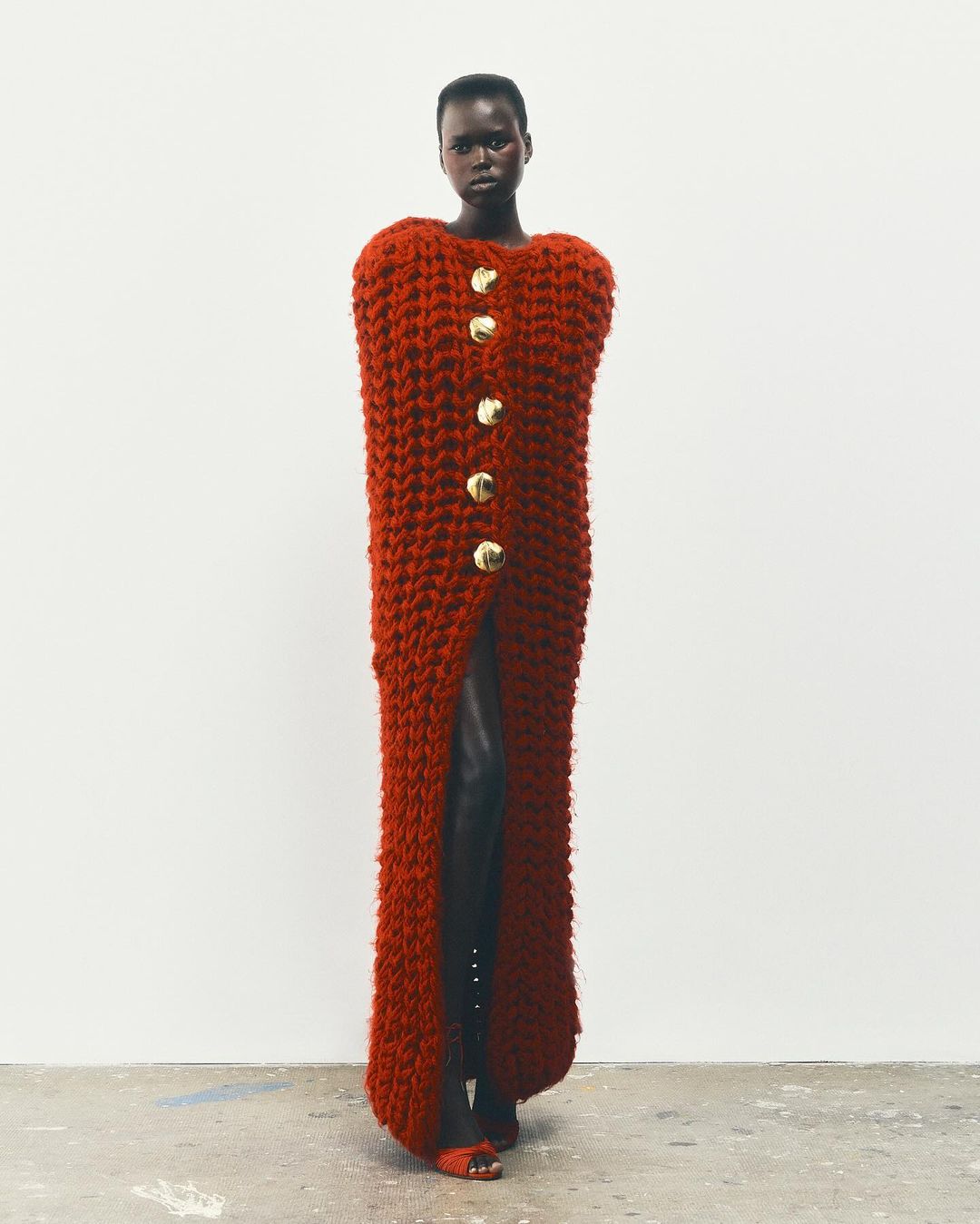 An African woman stands in a studio wearing a long, rust-colored knit dress with large gold buttons and dark heeled shoes, against a white backdrop.
