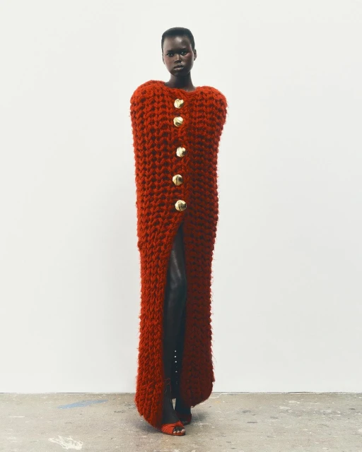 An African woman stands in a studio wearing a long, rust-colored knit dress with large gold buttons and dark heeled shoes, against a white backdrop.