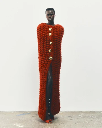 An African woman stands in a studio wearing a long, rust-colored knit dress with large gold buttons and dark heeled shoes, against a white backdrop.