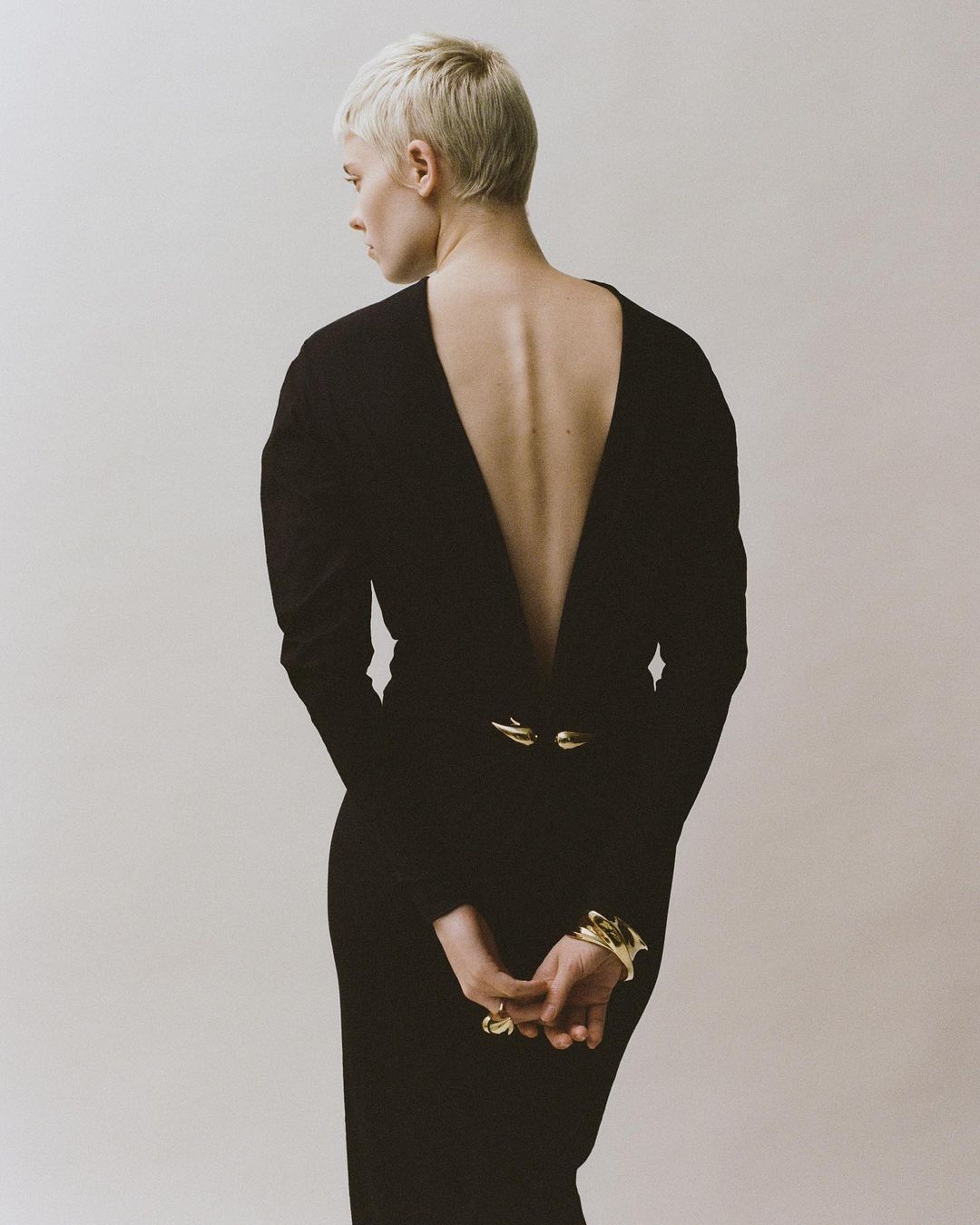 Blonde woman with short hair wearing a black long-sleeve dress with a deep V-back and gold jewelry, shot from behind in Paris.