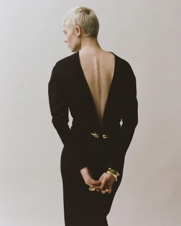 Blonde woman with short hair wearing a black long-sleeve dress with a deep V-back and gold jewelry, shot from behind in Paris.
