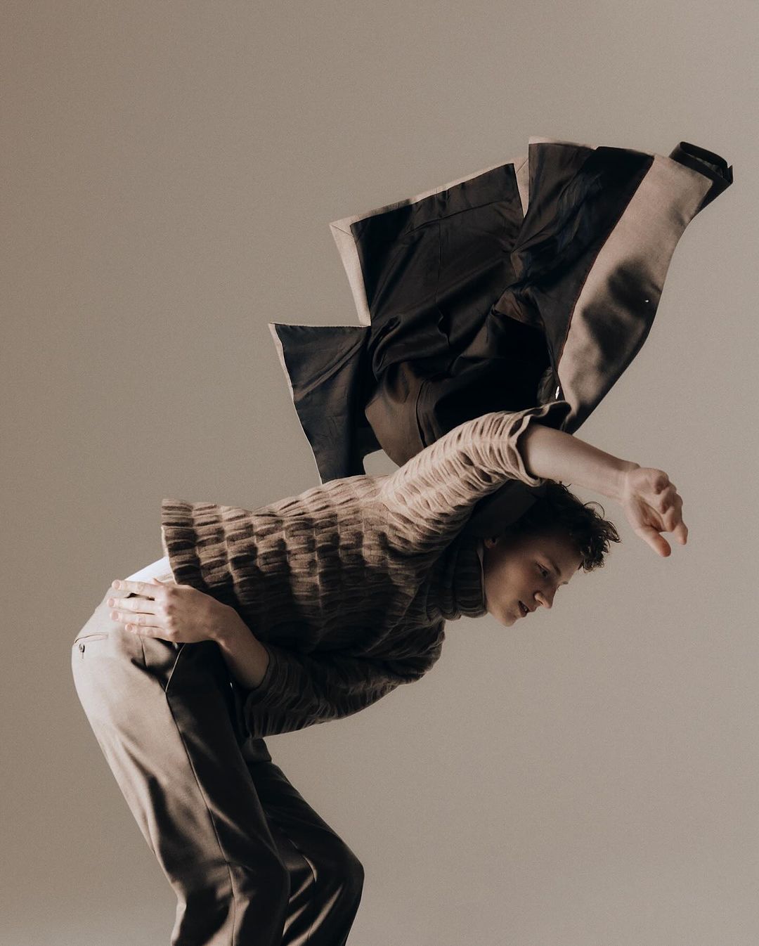 Fashion shot of a person in a sweater and trousers, leaning back with a jacket draped over their back, set against a neutral backdrop.