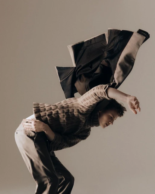 Fashion shot of a person in a sweater and trousers, leaning back with a jacket draped over their back, set against a neutral backdrop.