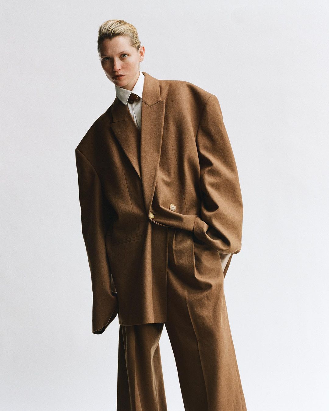 Woman in oversized brown suit and white shirt in a studio. Fashion editorial shot with clean white backdrop, full body portrait, and neutral tones.