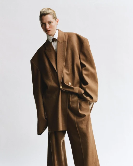 Woman in oversized brown suit and white shirt in a studio. Fashion editorial shot with clean white backdrop, full body portrait, and neutral tones.