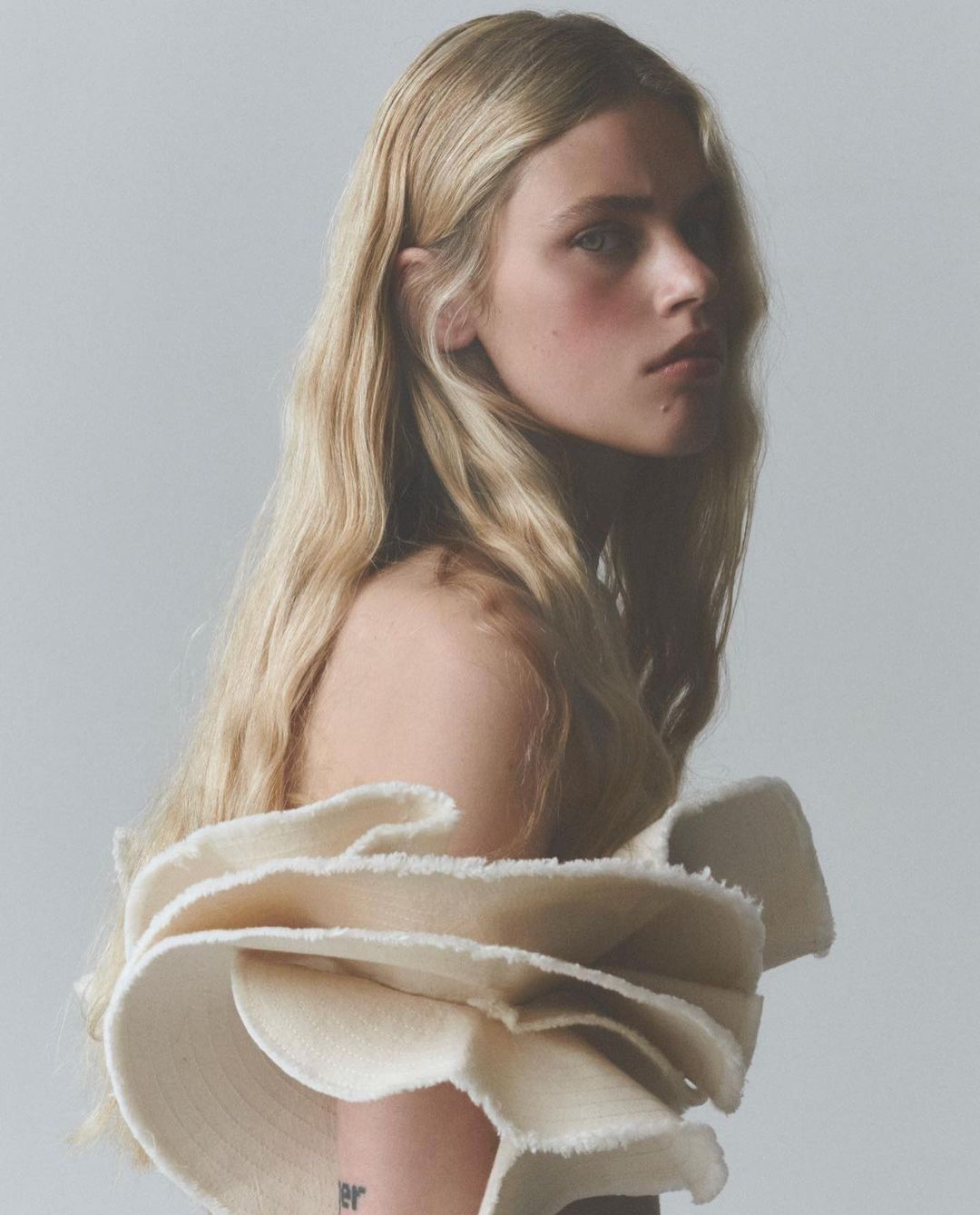 Portrait of a blond female model with pale skin wearing a ruffled off-the-shoulder garment against a pale background in a photo studio.