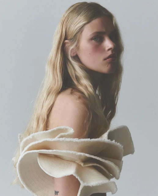 Portrait of a blond female model with pale skin wearing a ruffled off-the-shoulder garment against a pale background in a photo studio.