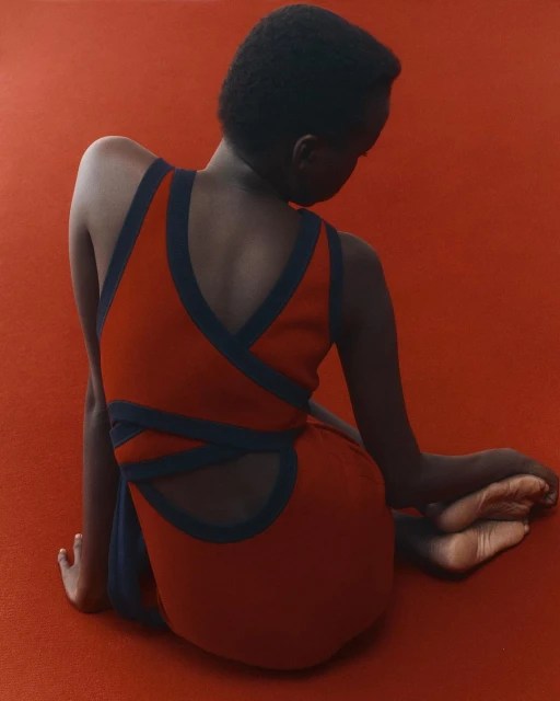 Woman with short dark hair wearing a red and dark blue dress is sitting on a matching red carpet with her back to the camera.