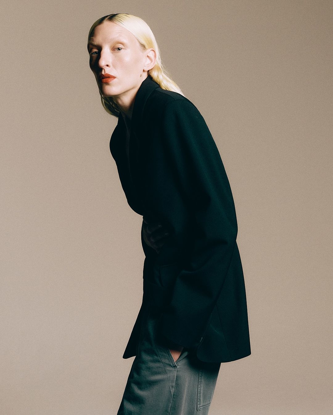 Model with blonde hair wearing a black blazer and grey trousers against a neutral background