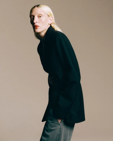 Model with blonde hair wearing a black blazer and grey trousers against a neutral background