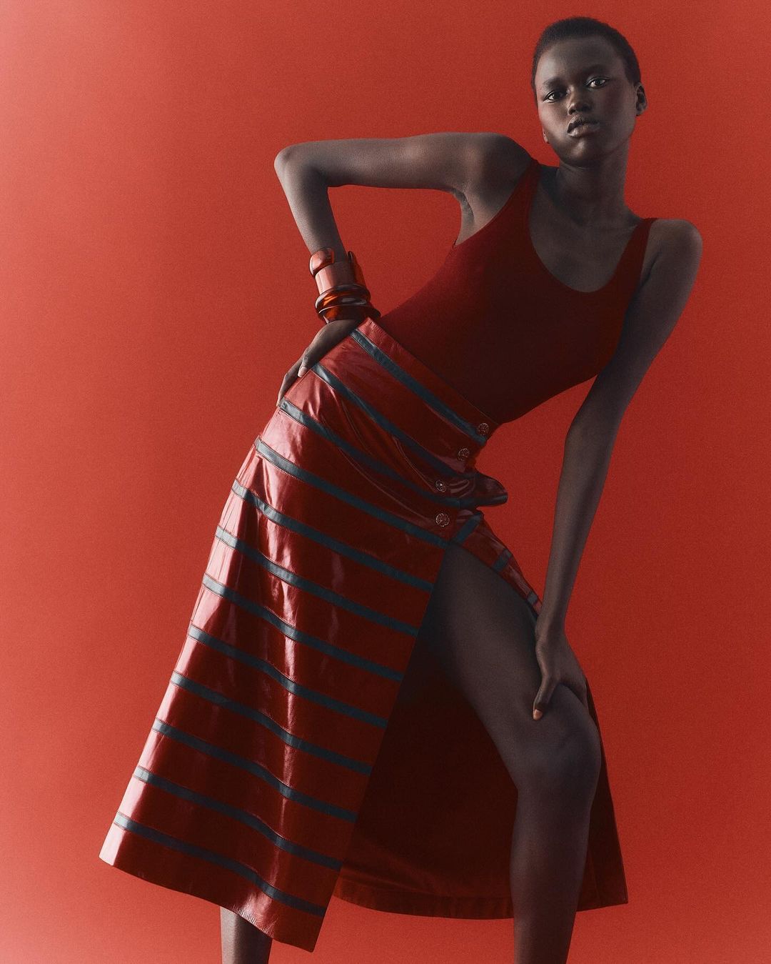 Fashion editorial shot of an adult woman posing in a red tank top and striped skirt against a red background in a studio.