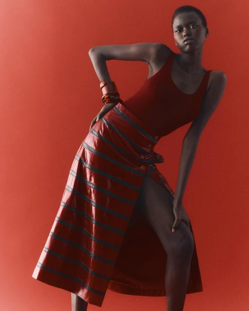 Fashion editorial shot of an adult woman posing in a red tank top and striped skirt against a red background in a studio.