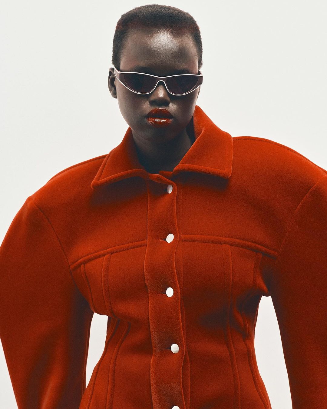 Model wearing dark futuristic sunglasses and a structured red jacket against a white background