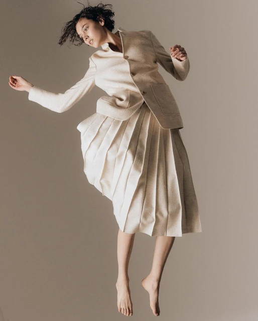 A barefoot woman in a cream-colored skirt suit poses mid-air against a neutral backdrop, captured in a fashion-forward, editorial style.
