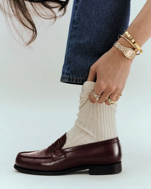 Burgundy leather loafer with beige knit socks, blue jeans, gold wristwatch, and gold bangle bracelet against a white backdrop.