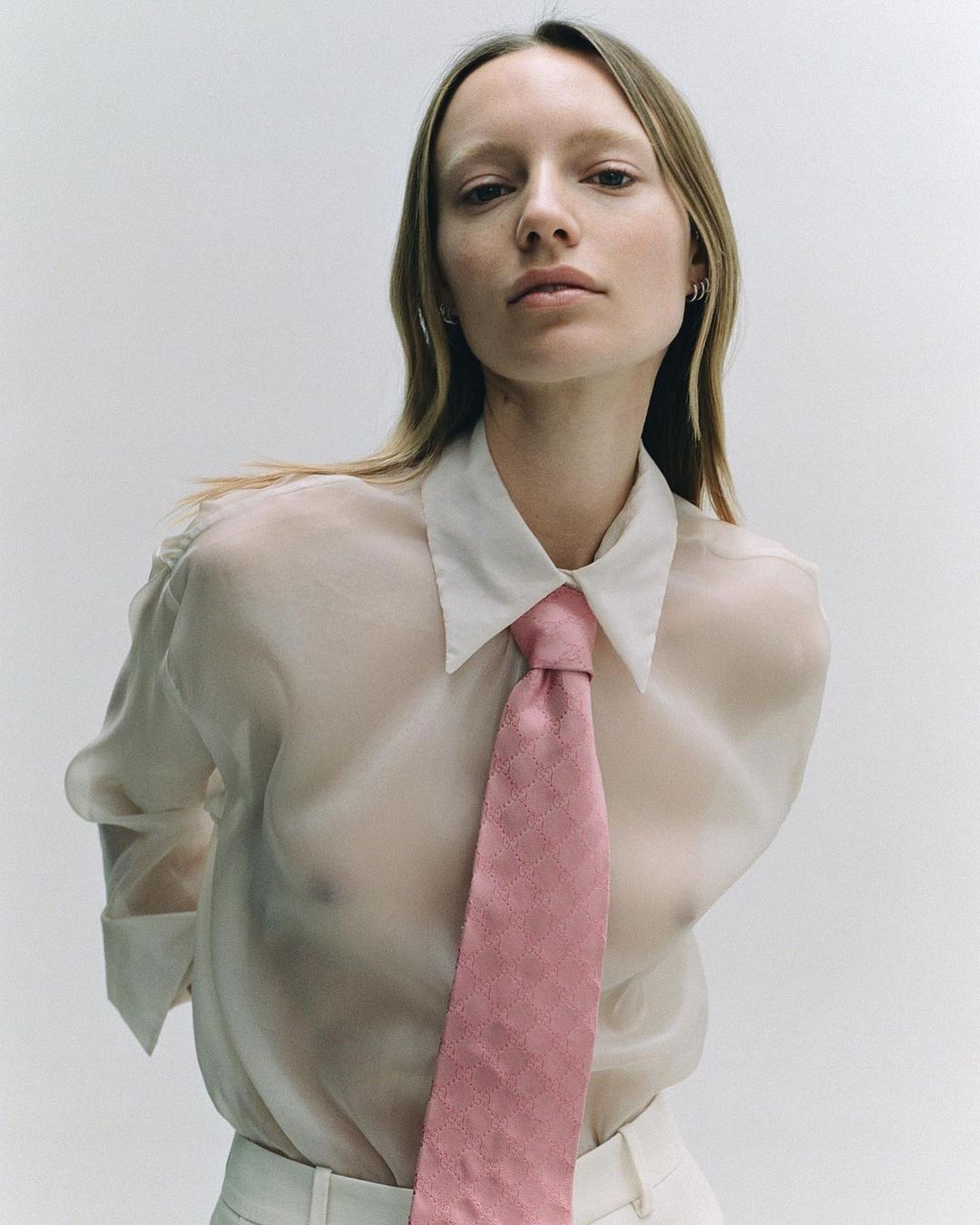Portrait of a fair-skinned woman wearing a semi-sheer blouse and pink necktie against a white backdrop in a studio setting.