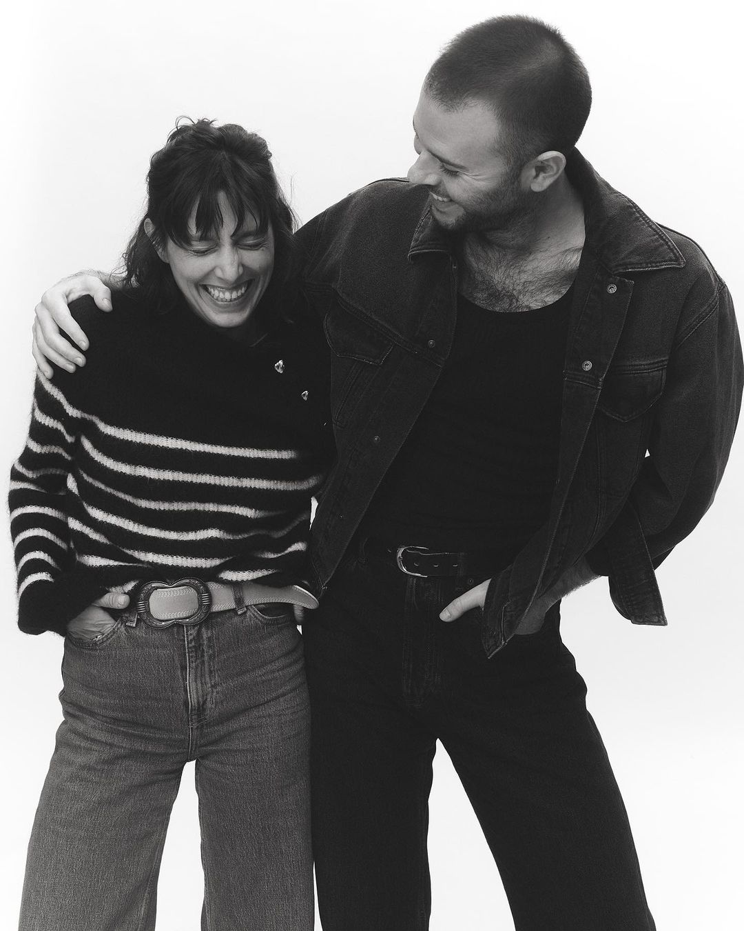 Black and white portrait of a smiling woman and man standing together against a seamless background, photographed by Anna Daki in Paris.