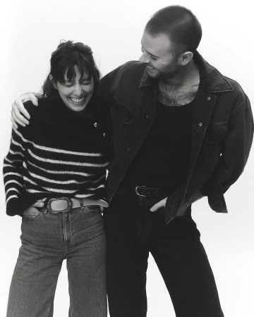 Black and white portrait of a smiling woman and man standing together against a seamless background, photographed by Anna Daki in Paris.