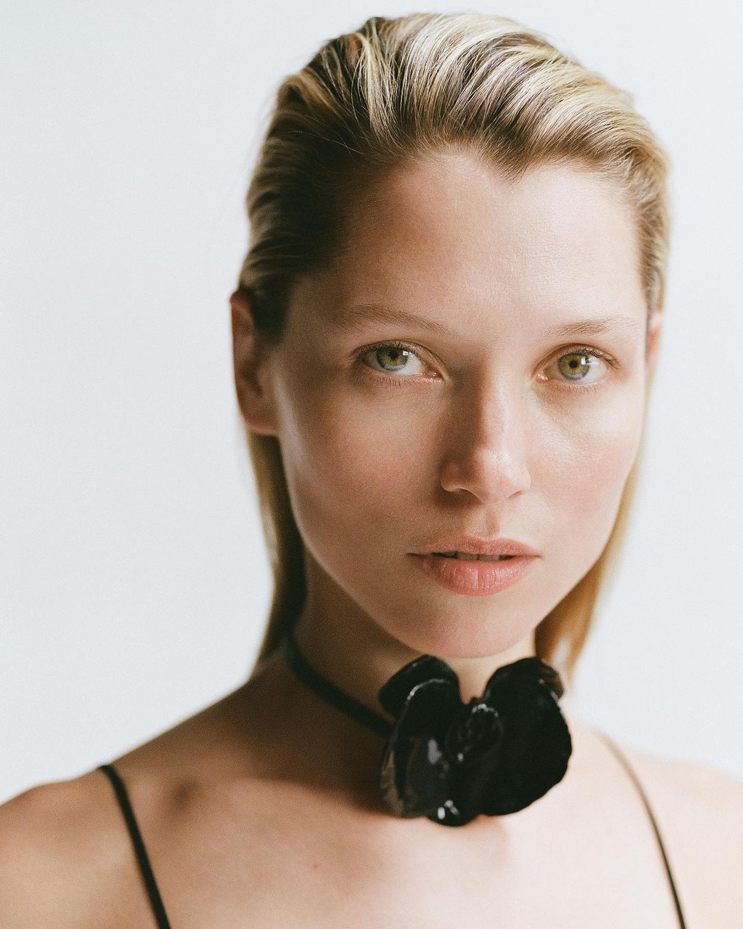 Close-up of a blonde woman with fair skin, wearing a black floral choker necklace, against a plain white studio background.