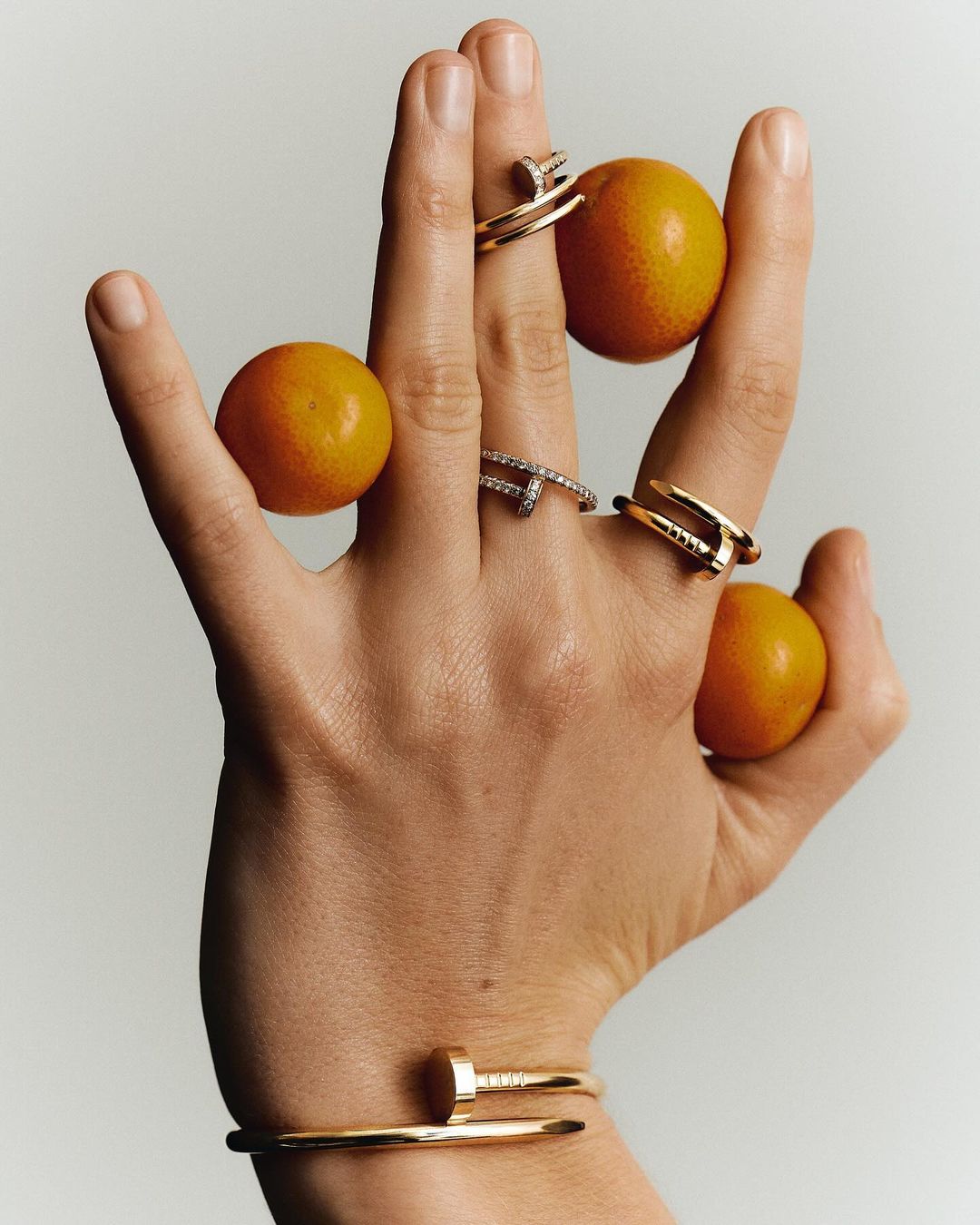 Close-up of a hand adorned with nail-inspired rings and a bracelet, holding tangerines against a neutral backdrop. Jewelry theme.