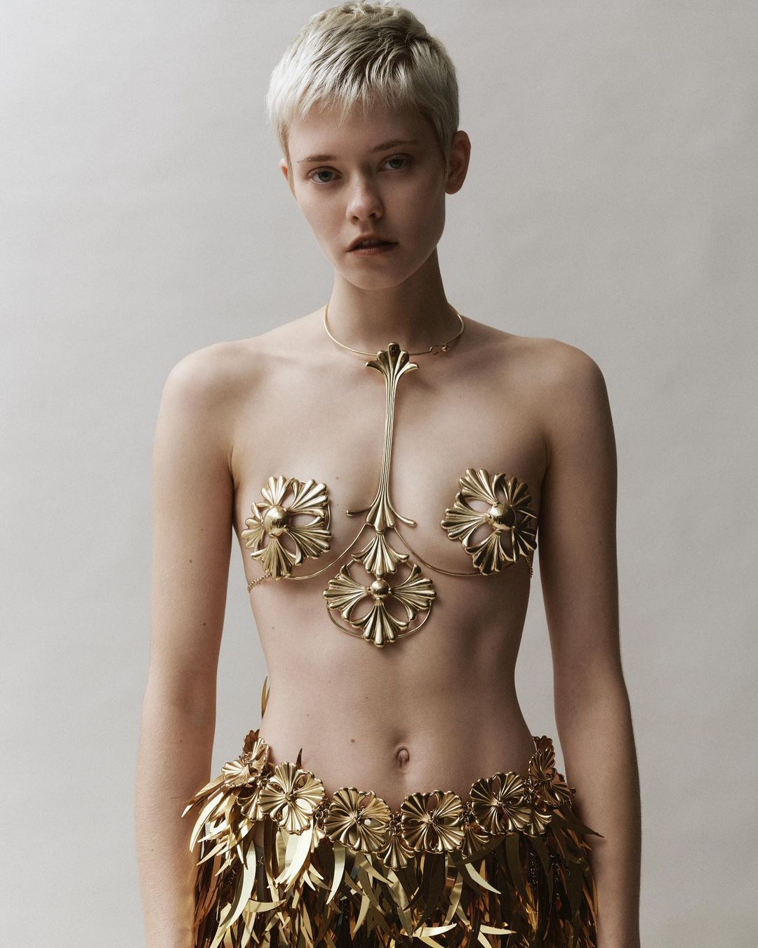 A young woman with short hair poses in an indoor studio, wearing ornate gold body jewelry and a feathered skirt against a neutral backdrop.