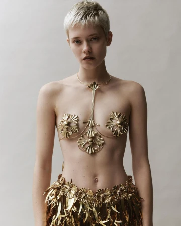 A young woman with short hair poses in an indoor studio, wearing ornate gold body jewelry and a feathered skirt against a neutral backdrop.