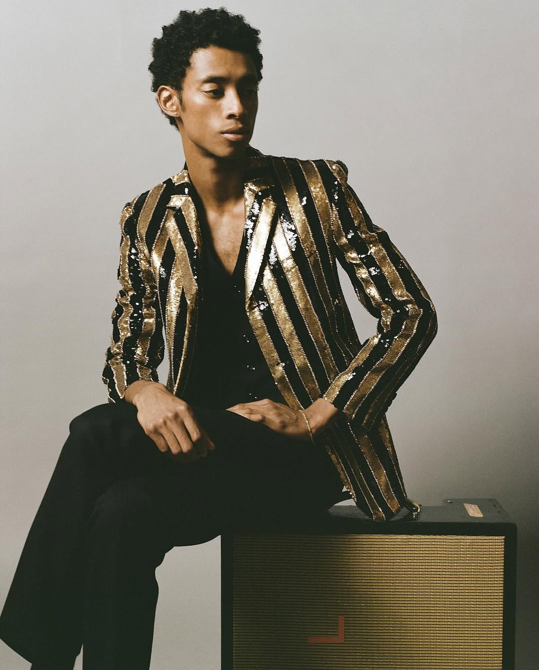 Portrait of a young adult male model seated on a speaker, wearing a black shirt and trousers with a gold and black striped blazer.
