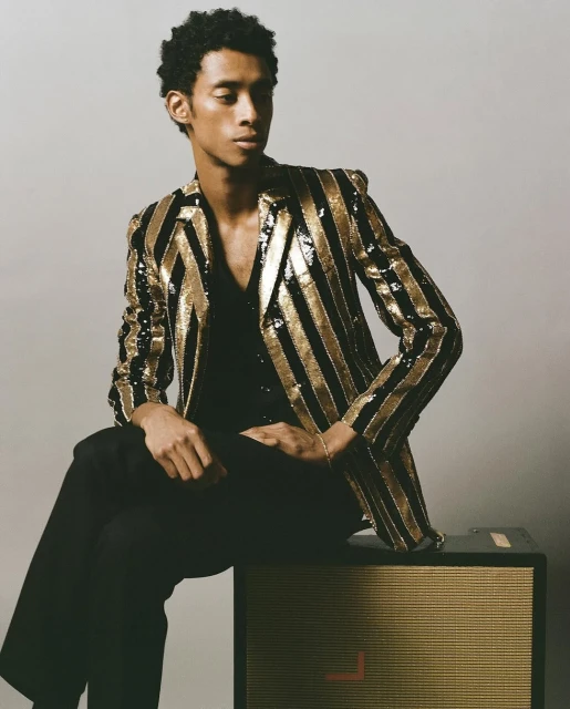 Portrait of a young adult male model seated on a speaker, wearing a black shirt and trousers with a gold and black striped blazer.