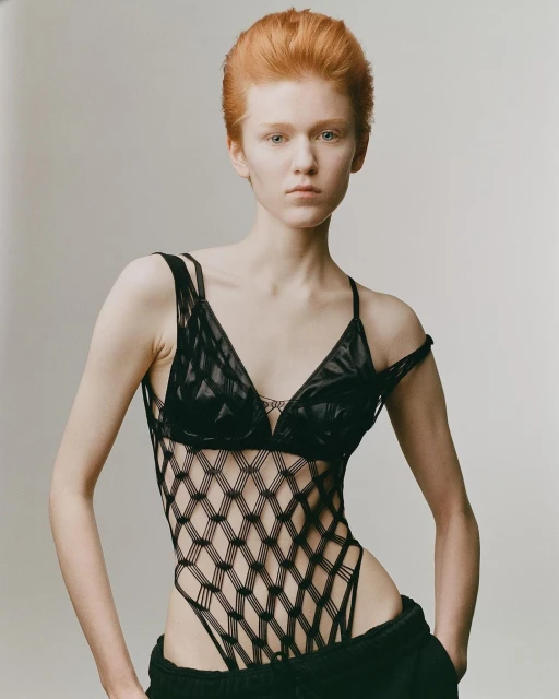 A redhead woman in a fashion bodysuit poses in a studio, eye-level angle, neutral facial expression, and soft lighting on a neutral background.