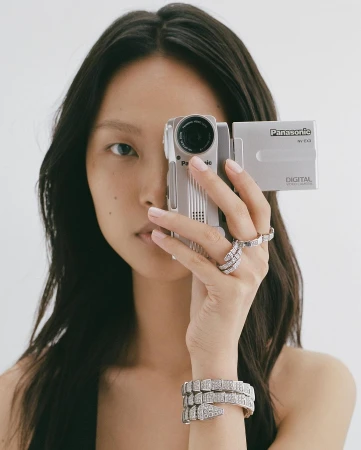 Young woman holds a vintage Panasonic video camera with her manicured hand, wearing diamond rings and bracelet against a white background.