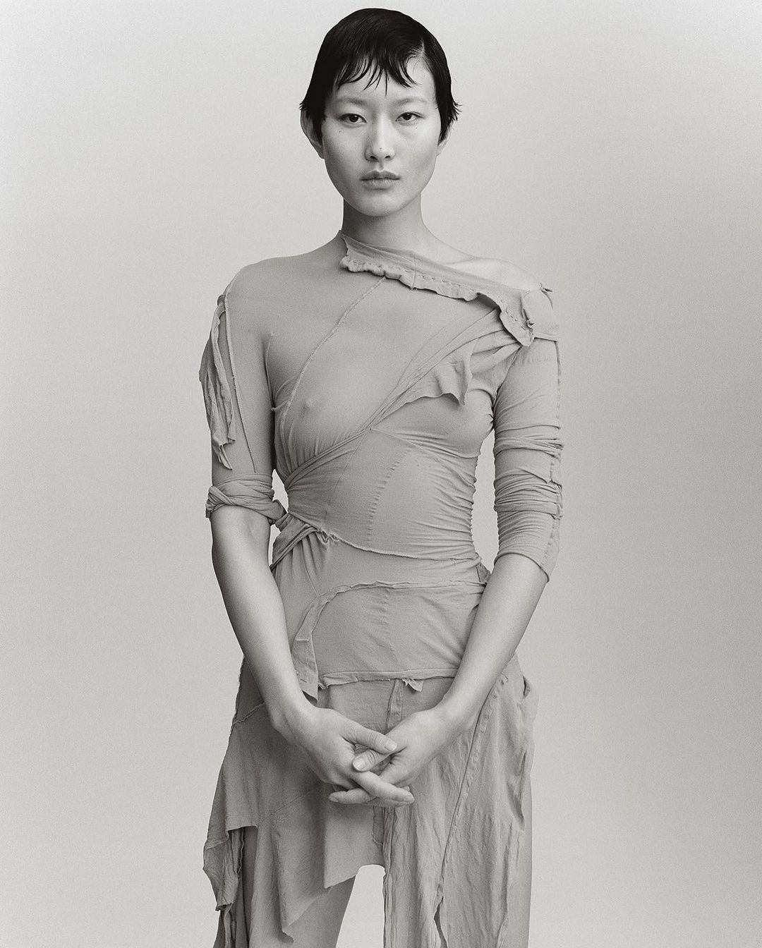 Monochrome portrait of a young Asian woman in a sheer, draped garment against a bright neutral background. Fashion editorial.