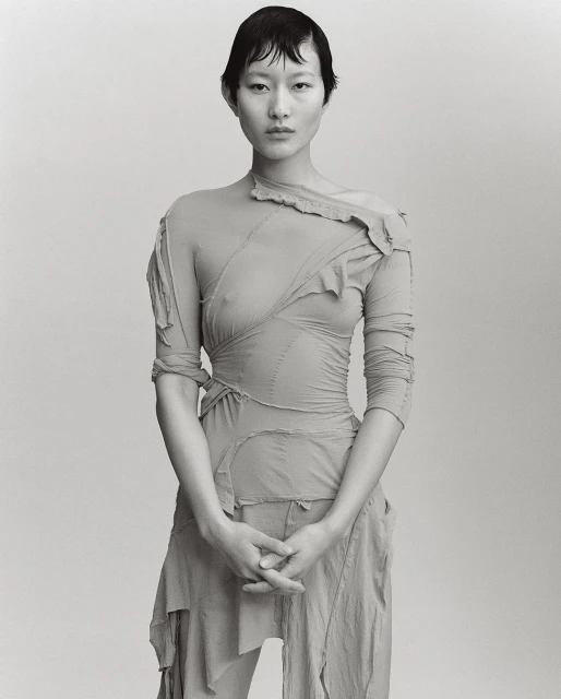 Monochrome portrait of a young Asian woman in a sheer, draped garment against a bright neutral background. Fashion editorial.
