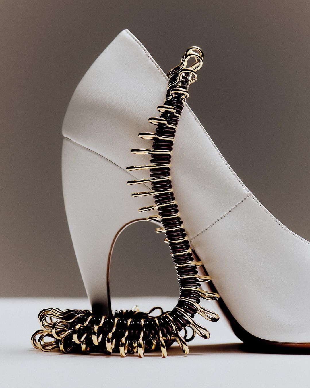 Elegant white high heel pump adorned with a gold and black coil jewelry piece, set against a neutral background in a studio setting.