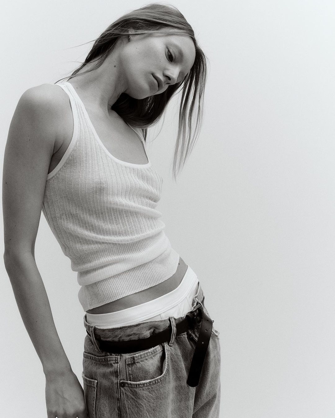 Young woman in a white ribbed tank top and blue jeans poses in a studio. The high-key monochromatic image focuses on fashion and portraiture.