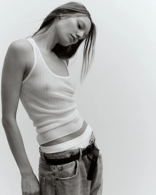 Young woman in a white ribbed tank top and blue jeans poses in a studio. The high-key monochromatic image focuses on fashion and portraiture.