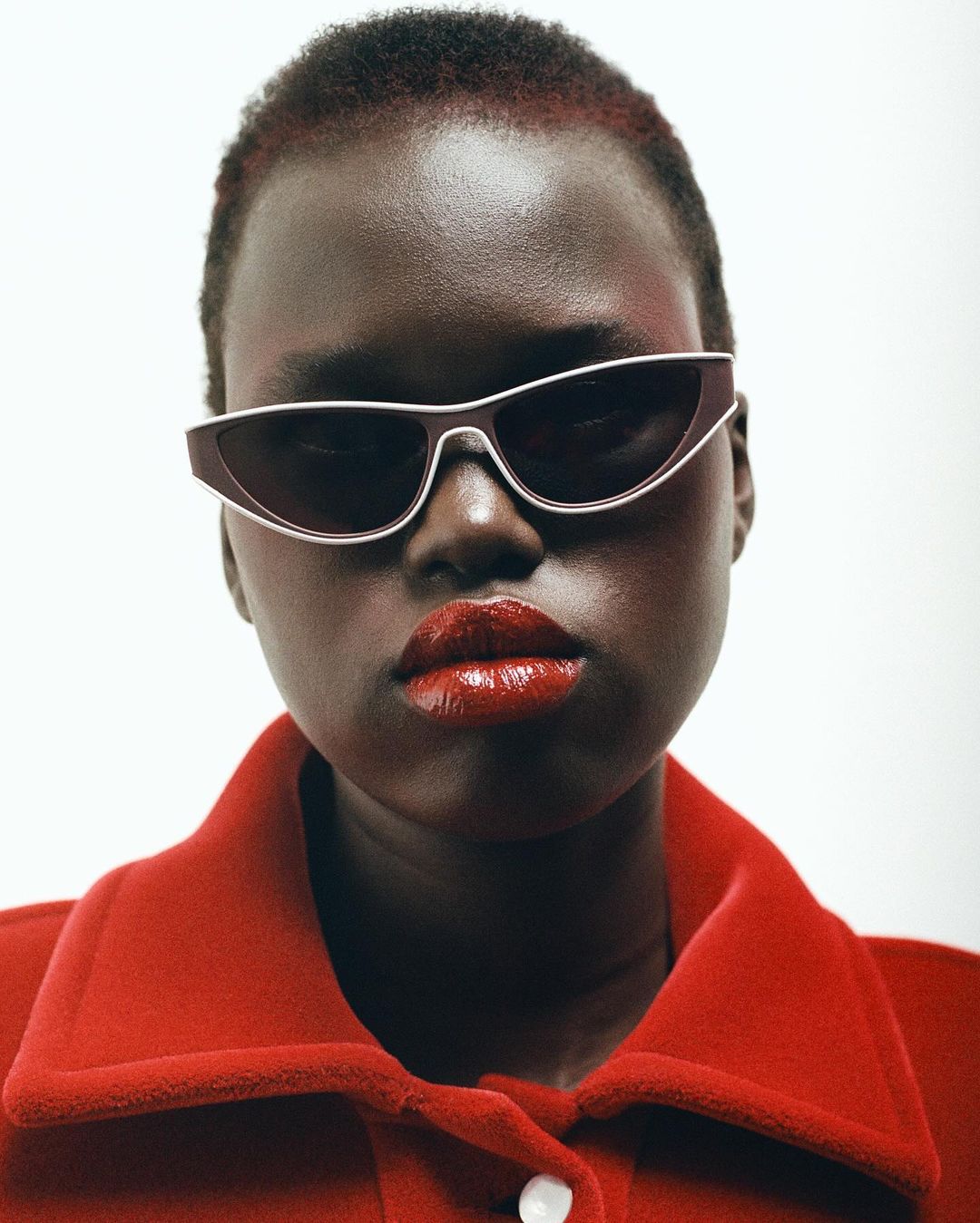 Close-up portrait of a dark-skinned woman with short hair wearing sunglasses and a red jacket, isolated against a white background.