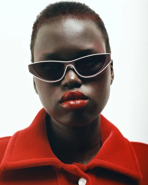 Close-up portrait of a dark-skinned woman with short hair wearing sunglasses and a red jacket, isolated against a white background.