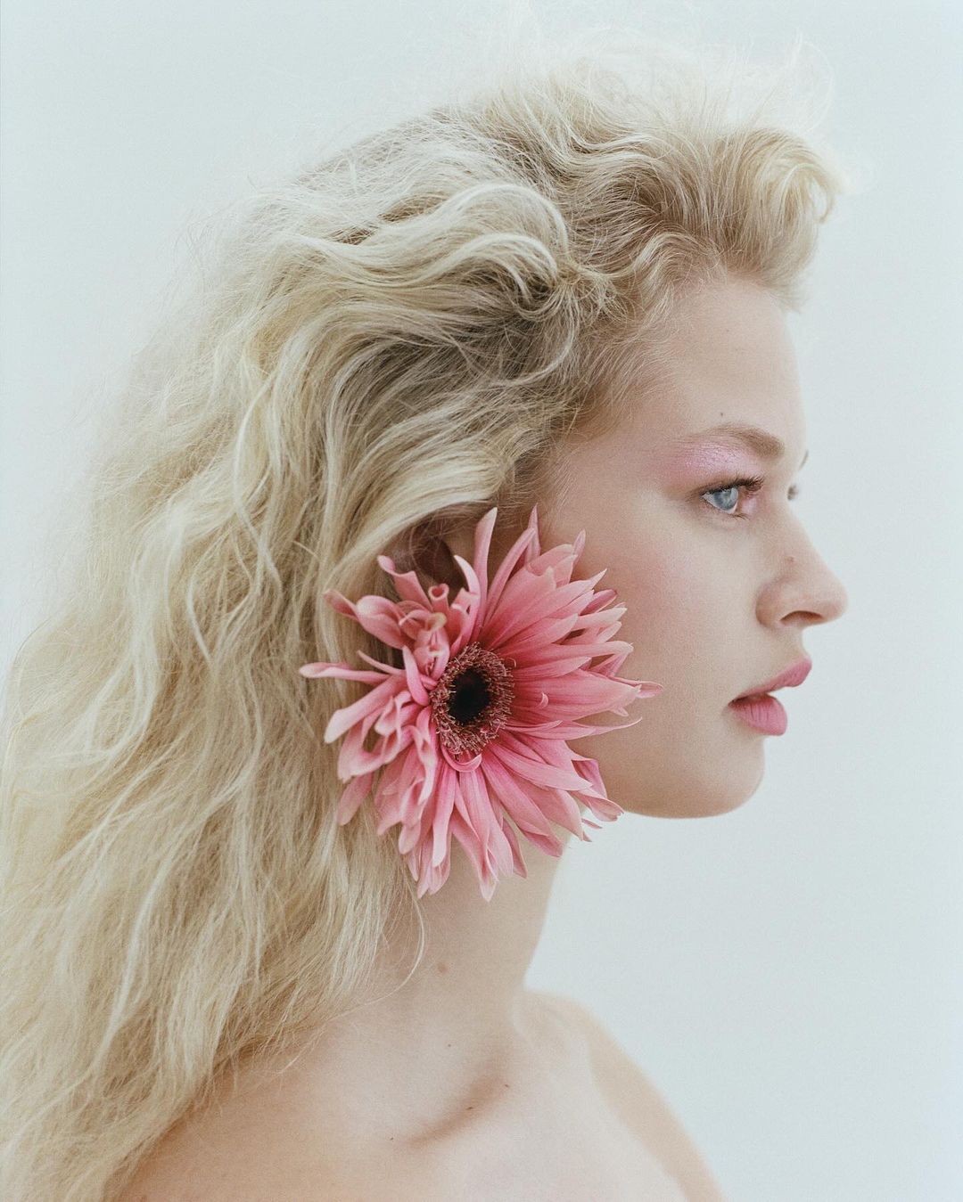 Profile view of a blonde woman with pink eye makeup, wearing a pink flower as a hair accessory, against a white backdrop.
