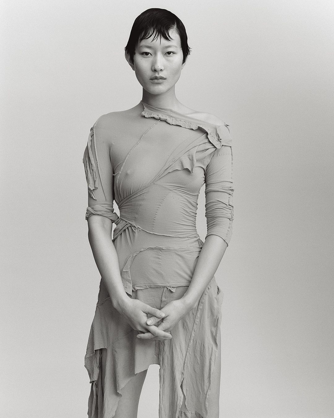 Monochromatic studio portrait of a slender Asian woman with short dark hair, wearing a pale draped dress with one long sleeve and her hands folded.