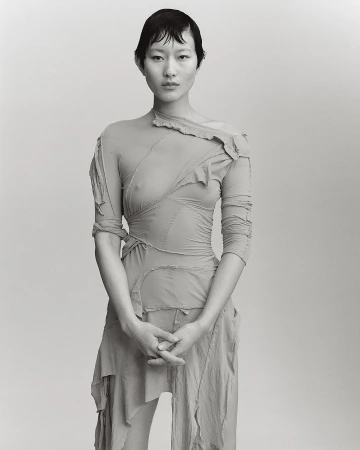Monochromatic studio portrait of a slender Asian woman with short dark hair, wearing a pale draped dress with one long sleeve and her hands folded.