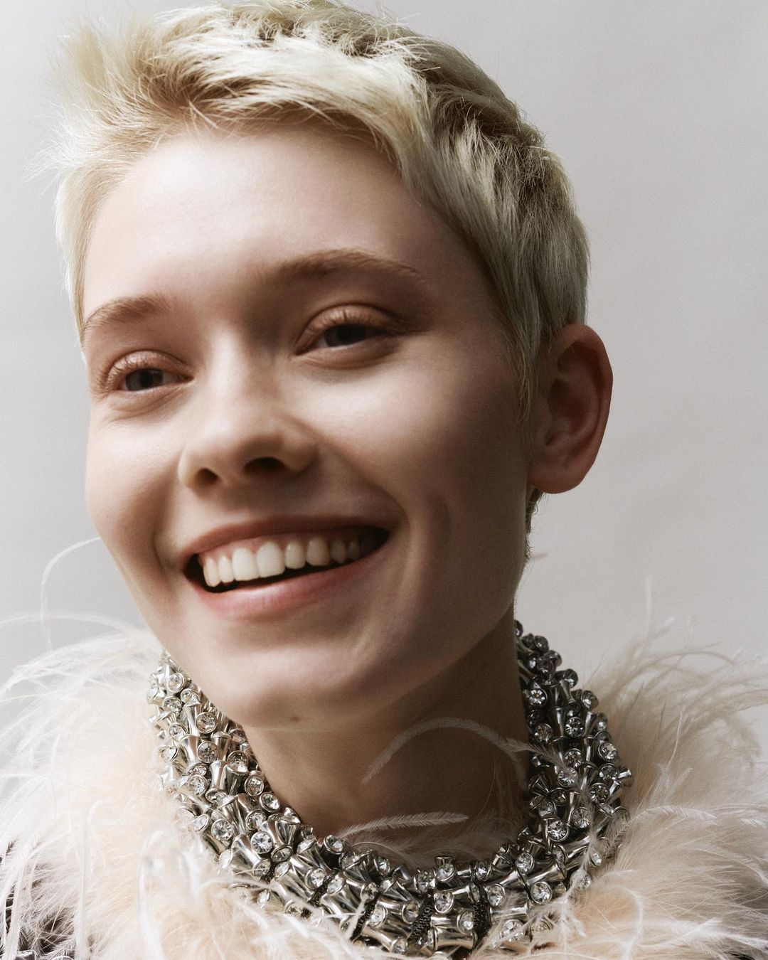 Close-up of a smiling adult woman with short blonde hair wearing a crystal and feather necklace against a plain grey background.