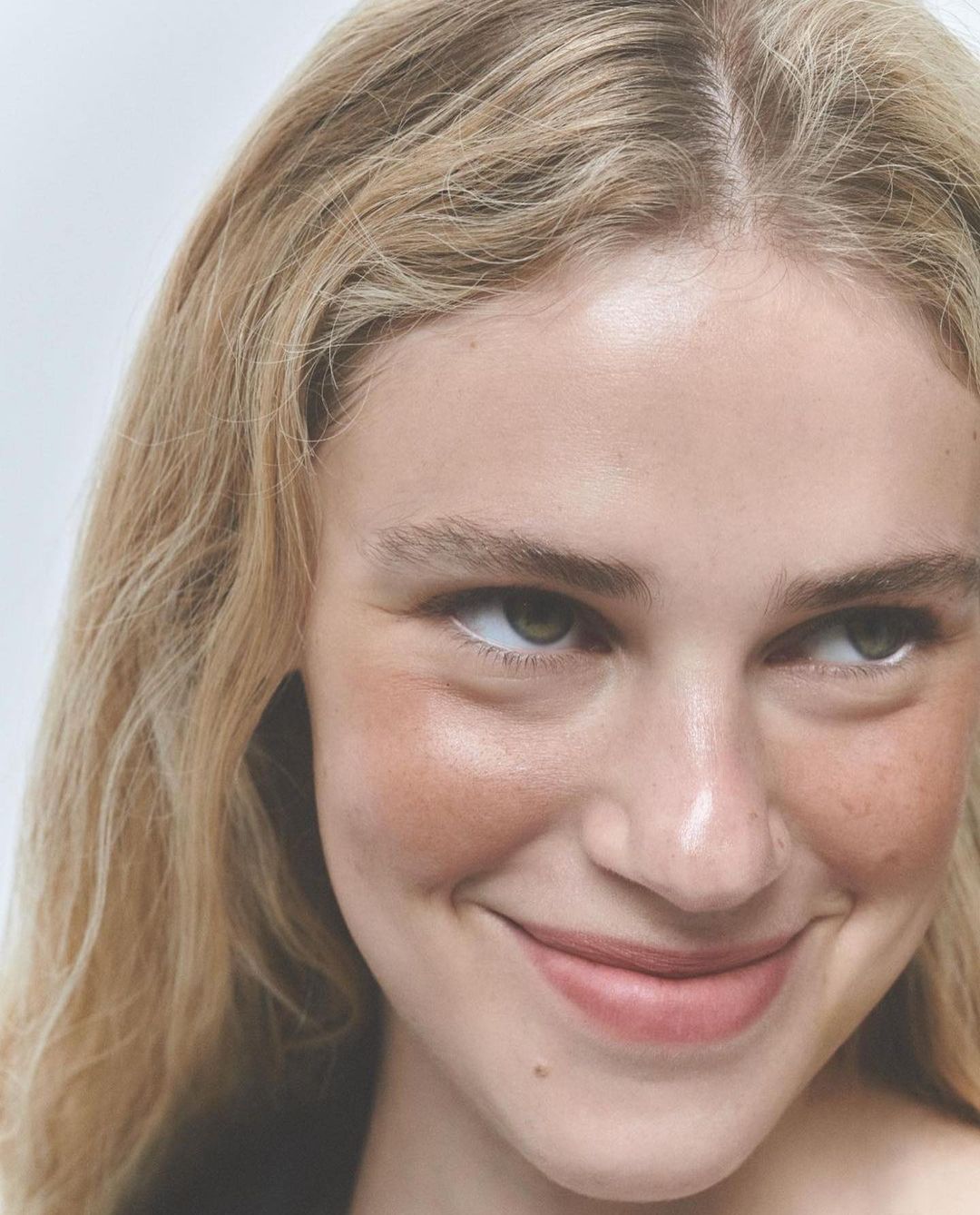 Close-up of a smiling young woman with blonde wavy hair. The portrait showcases her fair skin, light eye makeup, and a natural pink lip.