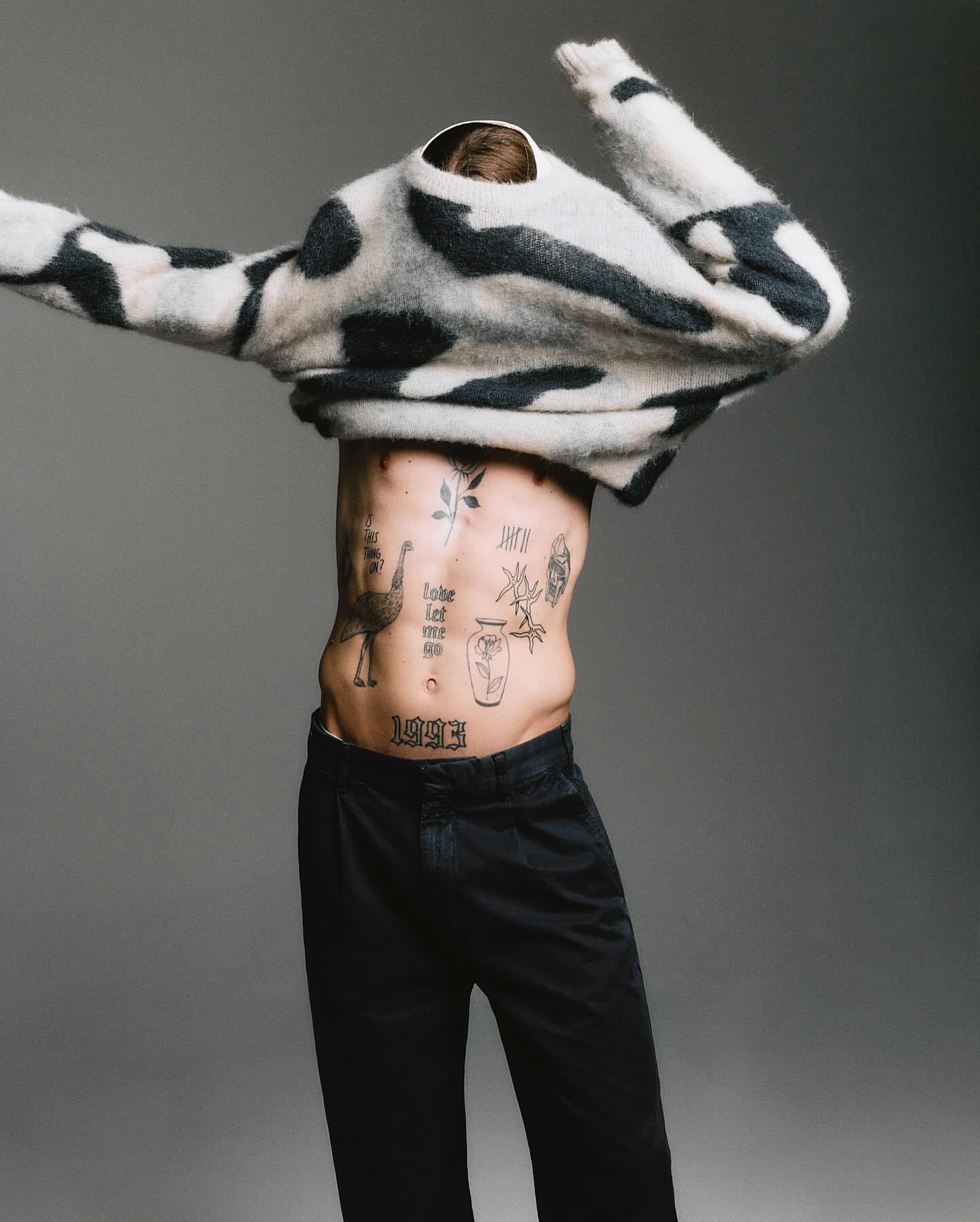 Shirtless man with torso tattoos pulling off a patterned monochrome sweater against a grey studio backdrop.